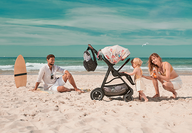 A man is sitting on the beach looking at a mother and child - the mother is spreading sunscreen on their child by the stroller