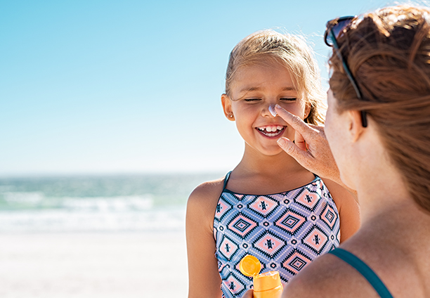 A baby girll stands forward smiling - mom applies sunscreen to her nose