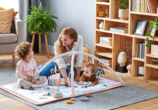 A woman is sitting on an educational mat with two children, laughing, the kids are playing